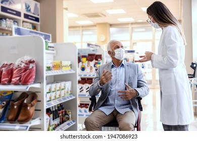 A Man In A Wheelchair Talks To A Female Pharmacist In White Uniform At The Pharmacy. Discussion Of Medical Therapy And Sale Of Drugs. They Both Wear A Protective Face Mask Due To The Corona Virus