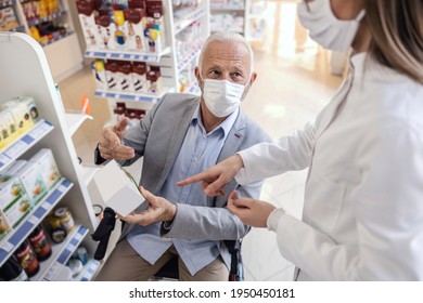 A Man In A Wheelchair Talks To A Female Pharmacist In White Uniform At The Pharmacy. Discussion Of Medical Therapy And Sale Of Drugs. They Both Wear A Protective Face Mask Due To The Corona Virus