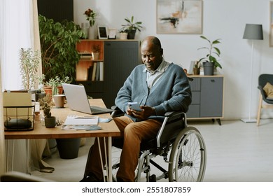 Man in wheelchair using smartphone while working from home office surrounded by books, plants, laptop, and desk setup in a bright, neatly organized room
