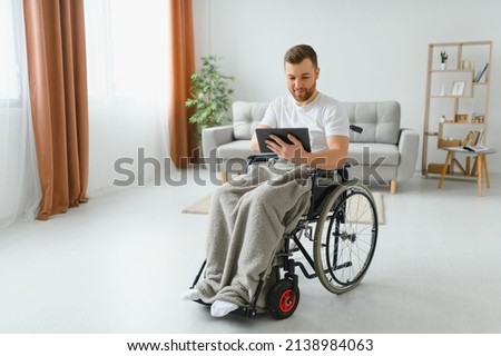 Man in wheelchair using tablet computer at home. Positive retired male with physical disability browsing web on touch pad, watching video online indoors