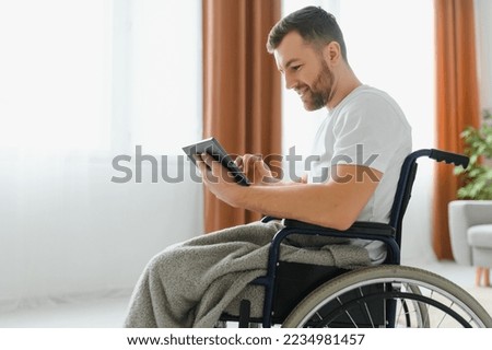 Man in wheelchair using tablet computer at home. Positive retired male Person with disability browsing web on touch pad, watching video online indoors
