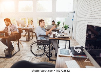 A Man In A Wheelchair Is Working In A Bright Office. His Colleagues Are Passionate About Work. The Man Looks Thoughtfully At The Screen.