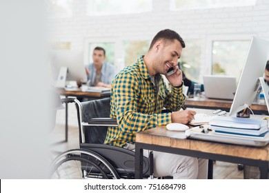 A Man In A Wheelchair Writes With A Pen In A Notebook And Talks On The Phone. He Is Working In A Bright Office And Smiling. His Colleagues Work Behind Him.