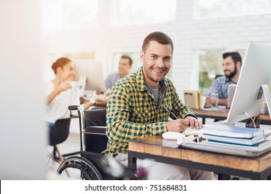 A Man In A Wheelchair Writes With A Pen In A Notebook. He Is Working In A Bright Office And Smiling. His Colleagues Work Behind Him.