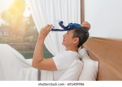 Man In White Clothes Laying With Knees Up In Bed  Hands Holding And Wearing CPAP Mask . Obstructive Sleep Apnea Therapy, Side View.