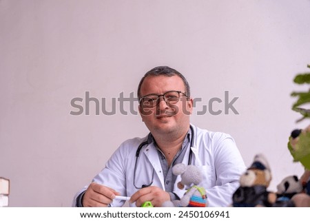 A man in a white coat is sitting at a desk with a stuffed animal on it. He is smiling and he is a doctor