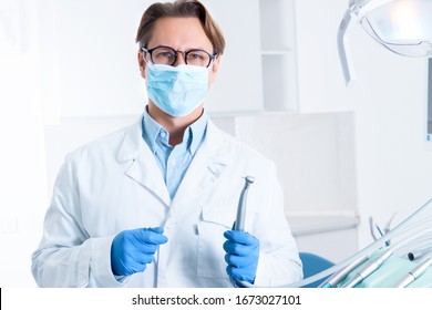 Man In White Doctor Robe Uniform, Glasses, Mask, Gloves Is Holding Instrument, Standing In Office, Clinic. Dentist Workplace. Orthodontist Is Preparing For Visit Of Patient. Dentistry Concept.