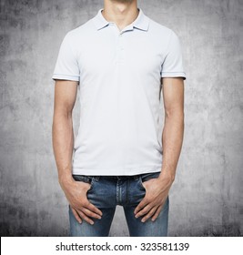 A Man In A White Polo Shirt And Denims Holds His Hands In Pockets. Concrete Background.