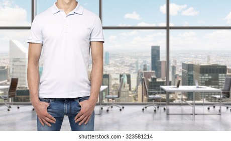 A Man In A White Polo Shirt And Denims Holds His Hands In Pockets. A Modern Panoramic Office Space In New York City. Manhattan Panoramic View.