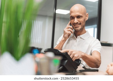 A man in a white polo shirt with a logo, smiling while talking on a phone at a desk. He is engaged in a conversation, with a plant and office items in the foreground. - Powered by Shutterstock