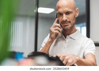 A man in a white polo shirt with a logo, talking on a phone in an office setting. He appears engaged and focused on the conversation, with a modern office background. - Powered by Shutterstock