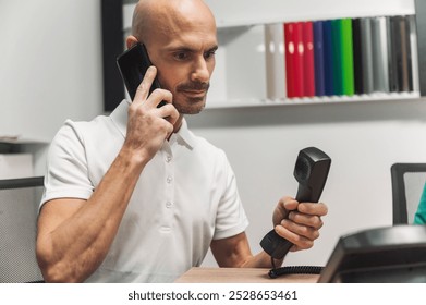 A man in a white polo shirt with a logo is engaged in a phone conversation, holding a smartphone in one hand and a traditional phone receiver in the other. He appears focused and is in an office setti - Powered by Shutterstock