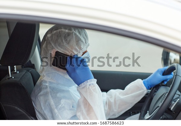 a man in a white protective medical suit with\
blue gloves, a mask in a car driving with a mobile phone during a\
coronavirus pandemic.\
horizontal