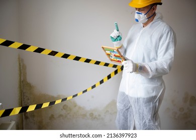 Man With White Protective Suit With Abacus Calculator, Slide Rule In Hand And Mouth Nose Mask In Front Of Yellow Black Barrier Tape In Front Of Wall With Mildew