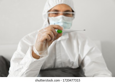 Man in white protective suit, medical mask, glasses and rubber gloves is holding in hand thermometer in a bed at his home. Epidemic, pandemic, coronavirus, 2019-ncov, covid-19, virus. - Powered by Shutterstock