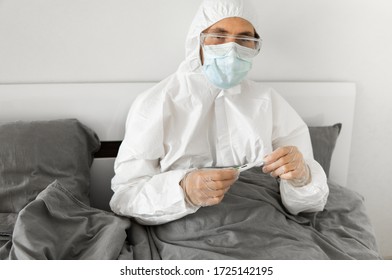 Man in white protective suit, medical mask, glasses and rubber gloves is holding in hand thermometer in a bed at his home. Epidemic, pandemic, coronavirus, 2019-ncov, covid-19, virus. - Powered by Shutterstock