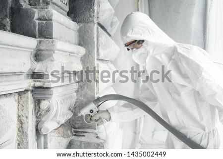 A man in a white protective uniform cleans stone carved sculpture from the dirt and concrete with a sandblasting machine. Restoration of stone sculpture. A jet of sand under high pressure.