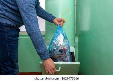 A man in white shirt and blue jean throwing black garbage bag in to plastic green trash.