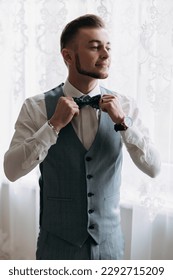 A man in a white shirt and a gray vest poses by the window and looks to the side, adjusts his blue bow tie. Front view. A stylish watch. Men's style. Fashion. Business