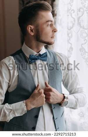 A man in a white shirt and a gray vest poses by the window and looks to the side, adjusts his vest. Front view. A stylish watch. Men's style. Fashion. Business