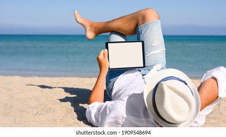 A Man In A White Shirt And Hat Is Lying On The Beach With A Tablet In His Hands. Blank White Display On Tablet.