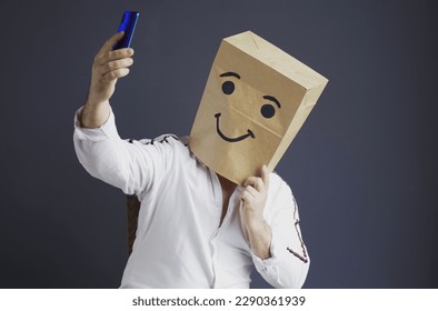 A man in a white shirt with a paper bag on his head, with a drawn smiley face, is talking on a video link or taking a selfie on a smartphone. - Powered by Shutterstock