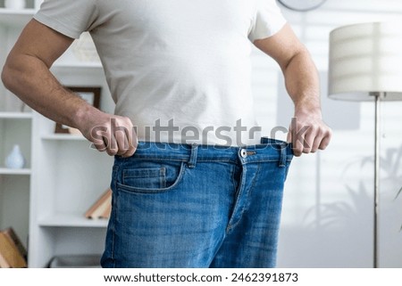 Man in a white shirt showcasing significant weight loss by wearing oversized jeans in a well-lit room.