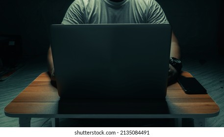 A man in a white shirt sits on a laptop in a dark room. with light shining down. internet concept addiction.topview.