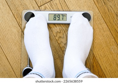 A man in white socks weighing himself on a transparent electronic floor scale, the weight reading is 89 kg, top view