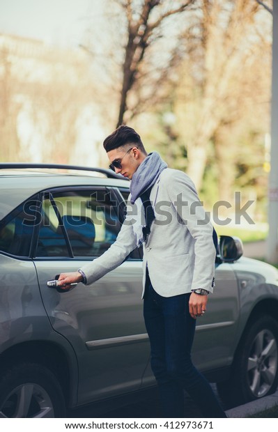 Man in a white suit and glasses waiting on the street\
near the car