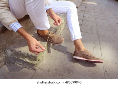 Man In White Trousers Sitting On The Steps In The City And Tying The Laces On The Shoes. Men's Summer Fashion, Stylish Men's Shoes. Brown Perforated Suede Summer Shoes With Laces