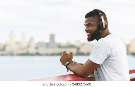 A man in a white t-shirt and headphones, leans on a bridge railing while listening to music and looking out at the city skyline. - Powered by Shutterstock