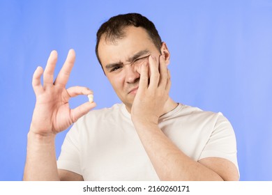 Man In A White T-shirt On A Blue Background Holds A Wisdom Tooth In His Hands After Surgical Tooth Extraction.Man After An Operation To Remove Wisdom Teeth.Pain In Wisdom Teeth, Concept Of Dentistry