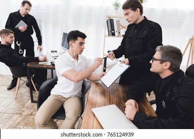 A Man In A White T-shirt Is Sitting In The Police Station. He's Talking To A Police Officer. They Talk About Crime. The Officer On The Desk Has A Paper.