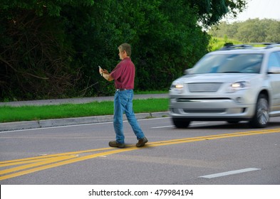 A Man Who Is Completely Distracted By His Cell Phone Is Unaware That He Is Jaywalking By Walking Out Into Heavy Traffic On A Busy Highway Road.