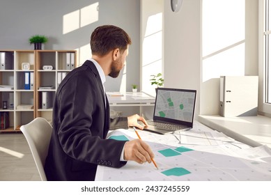 Man who is a professional corporate cadastral surveyor works with cartographic cadastre city maps, studies plot boundaries and numbers, uses digital graphic urban structure design plans on notebook PC