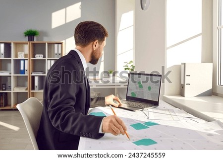 Man who is a professional corporate cadastral surveyor works with cartographic cadastre city maps, studies plot boundaries and numbers, uses digital graphic urban structure design plans on notebook PC