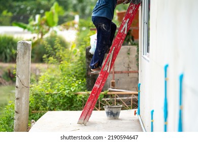 Man Who Wearing Dark Pants Is Climbing A Red Stairs To Fix The House. Green Shrub And Broken Pillar Are On The Background 