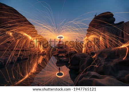 Man wielding rounded burning fire spark from steel wool in rock rapids of grand canyon reflection on river at Hat Chom Dao, Ubon Ratchathani
