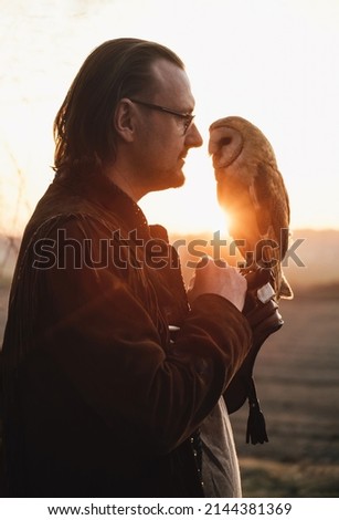 Man and wild bird over sunset sky in field looking on each other Owl symbol of power, wisdom wealth, learning person in eye glasses, vision problems. Nature and human partnership, friendship