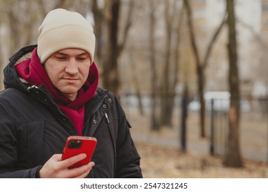 Man in winter attire checks smartphone outdoors in a park during autumn. - Powered by Shutterstock