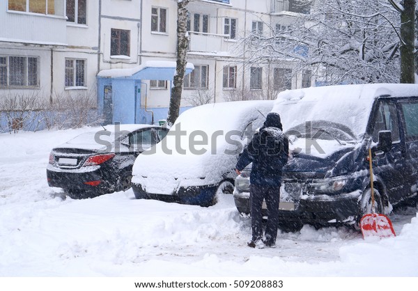 The man in\
the winter to clean the car from snow in heavy snow and blowing\
snow. Parking cars under a snow\
layer.