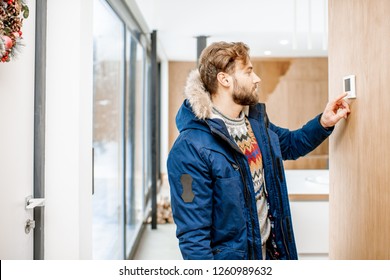 Man In Winter Clothes Feeling Cold Adjusting Room Temperature With Electronic Thermostat At Home