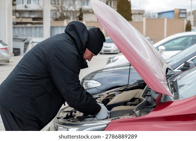 Man in winter jacket and hat checking car engine under open hood in parking lot on cloudy day. Car repair concept