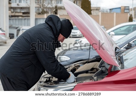 Man in winter jacket and hat checking car engine under open hood in parking lot on cloudy day. Car repair concept