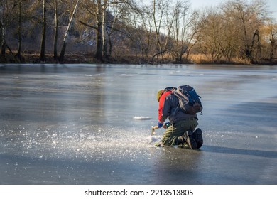 A man in a winter jacket, trousers and with a backpack and spinning nearbue diligently pierces the ice on the frozen river with an axe. Back view