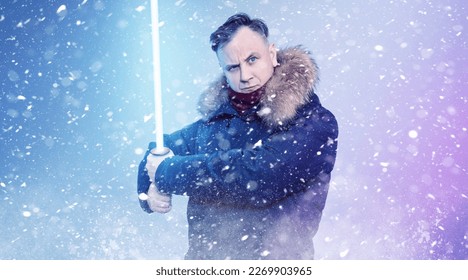 Man in a winter jacket without a hat with a light sword in his hands on a blue background with falling snow.

