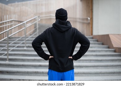 A Man In A Winter Running Uniform Stands In Front Of The Steps Up. Rear View Of Hooded Sportsman Before Run Upstairs.