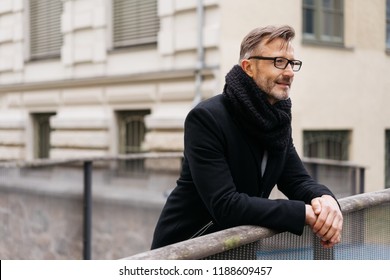 Man in a winter scarf and overcoat standing leaning on a bridge railing over a canal deep in thought staring straight ahead