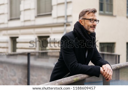 Man in a winter scarf and overcoat standing leaning on a bridge railing over a canal deep in thought staring straight ahead